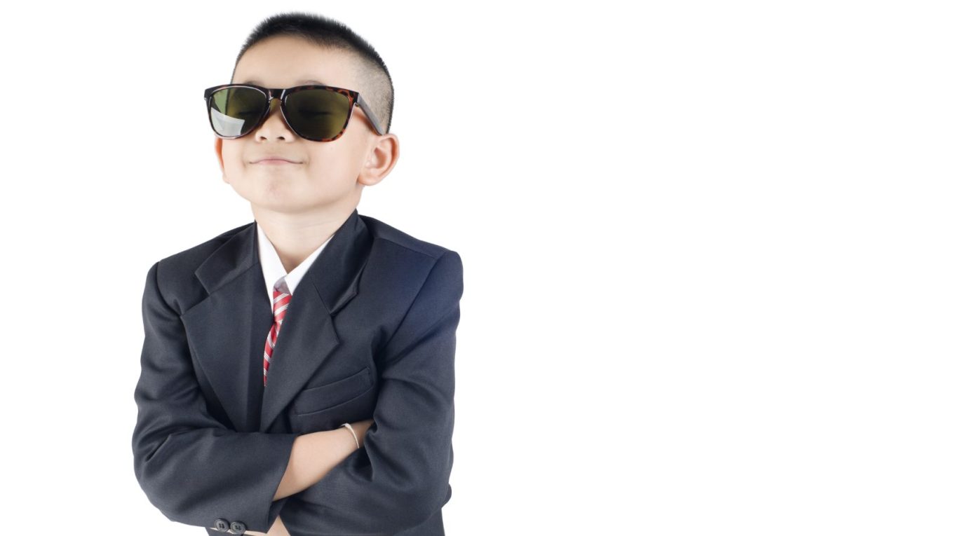 Ein Junge in einem Anzug und Sonnenbrille steht selbstbewusst mit verschränkten Armen.
